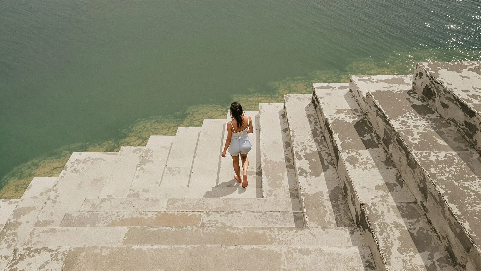 Woman walking barefoot down stone steps toward calm water with softened body posture