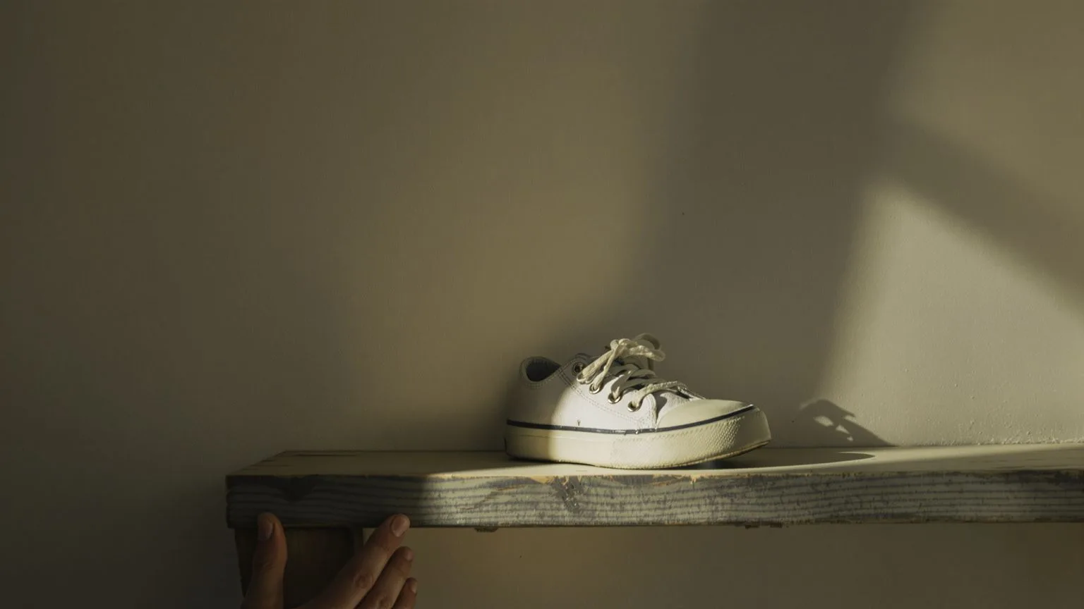 Adult hand reaching toward small worn child's shoe on weathered shelf in soft light, inner child healing symbol