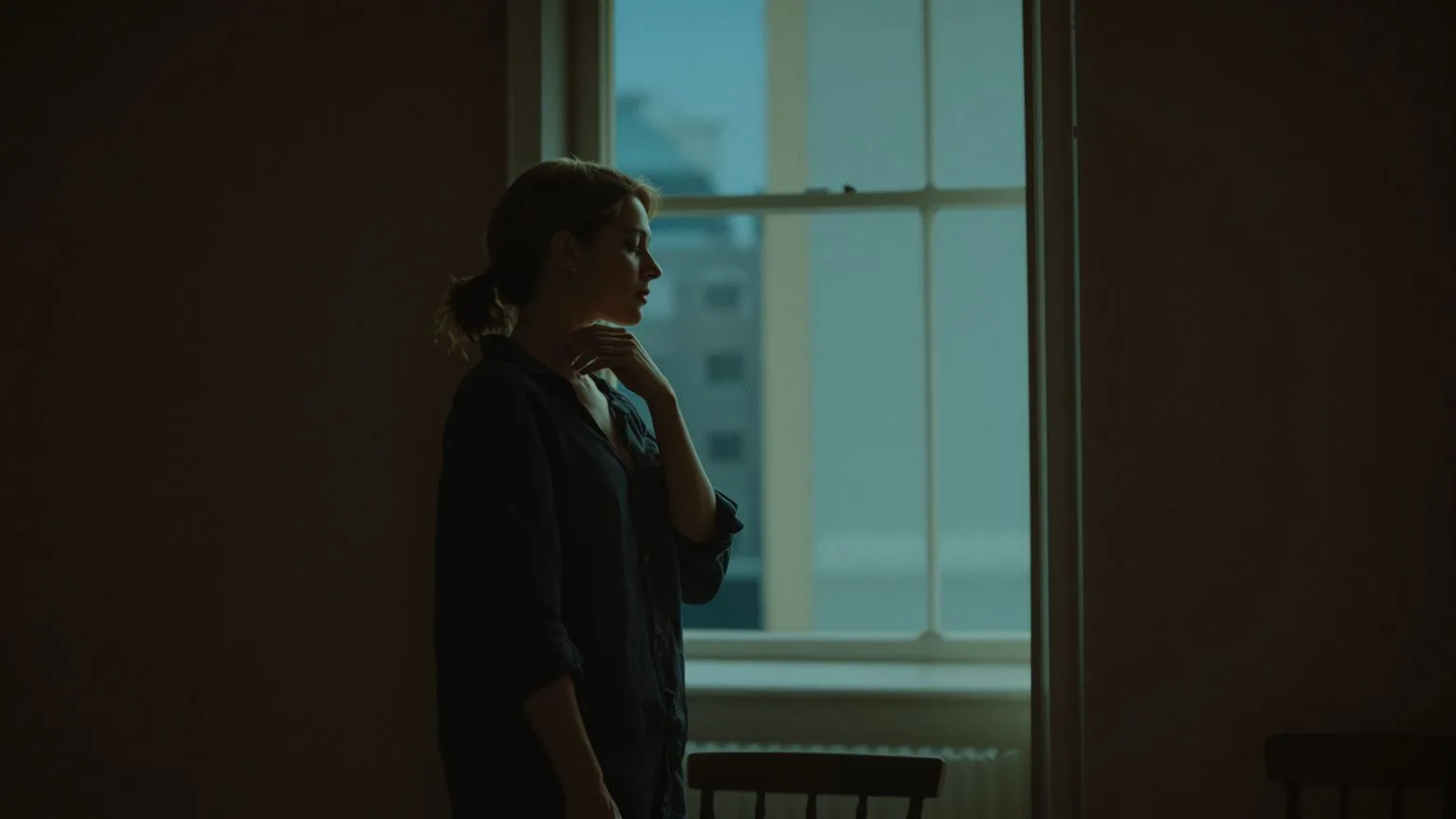 Woman touching her neck near a window at dusk, body slowing down before somatic exercises to release trauma free