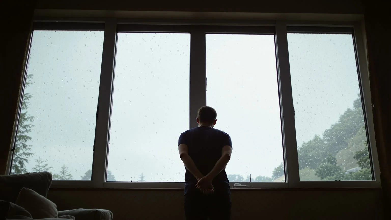 Person at window from behind, contemplating somatic yoga effects, hands clasped behind back