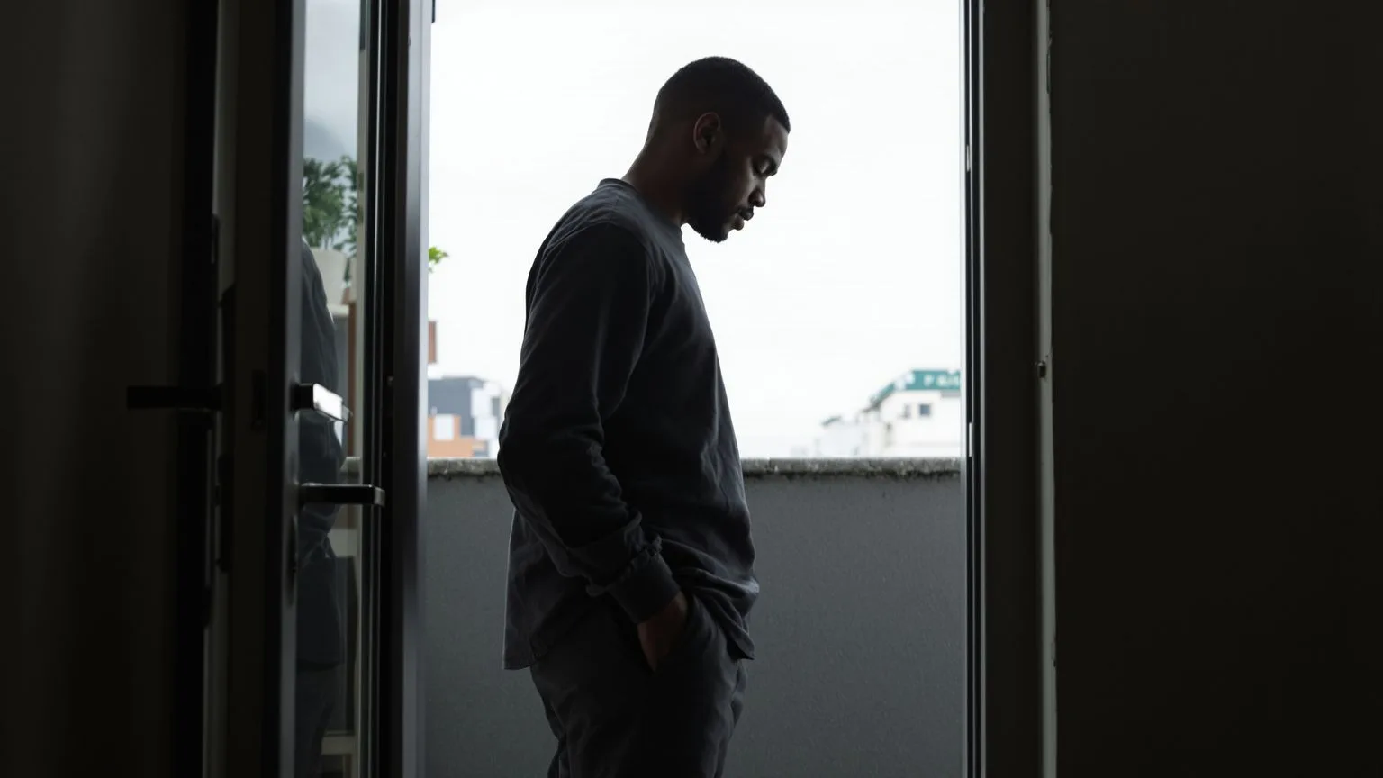 Man standing at balcony doorway threshold with body half in shadow showing the fear that drives the performance