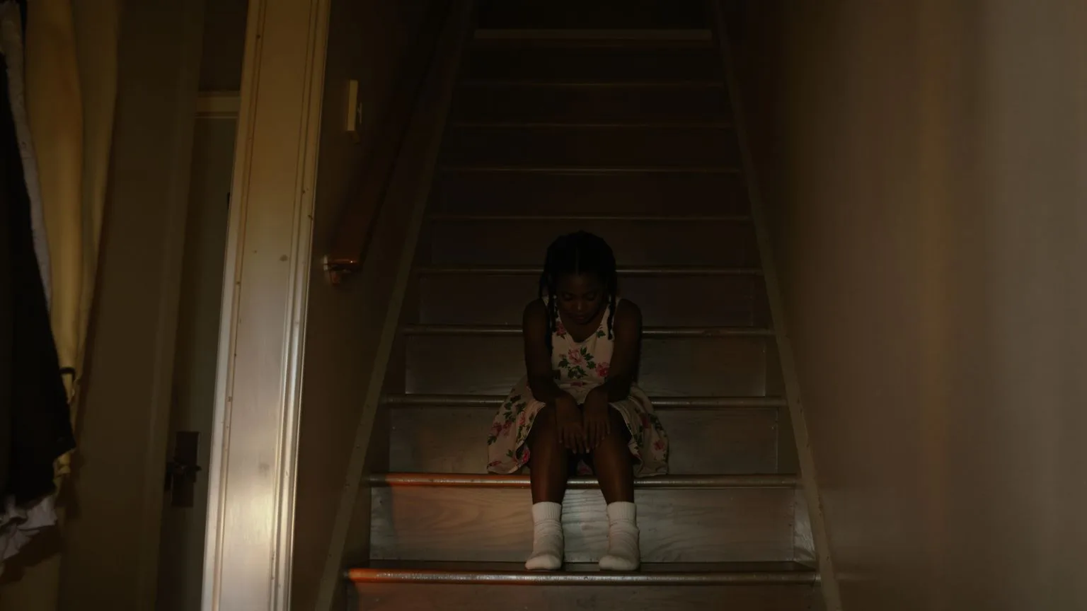 Young girl sitting alone on a dim staircase in warm doorway light, evoking where people pleasing was born in childhood