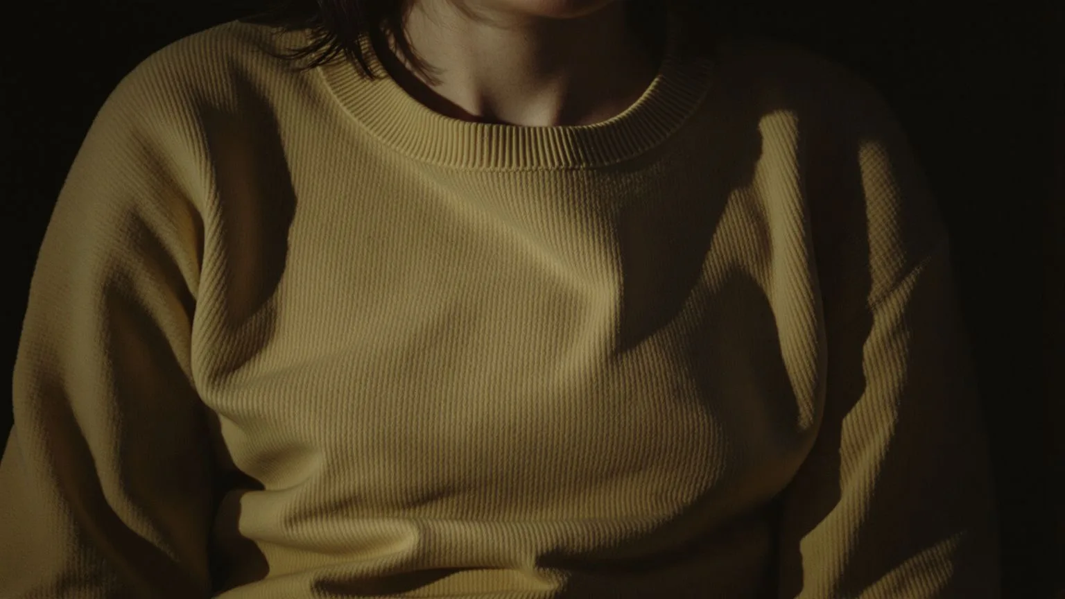 Close view of a woman's chest and collarbone curved inward in dim window light, quietly holding chest tightness anxiety in the body